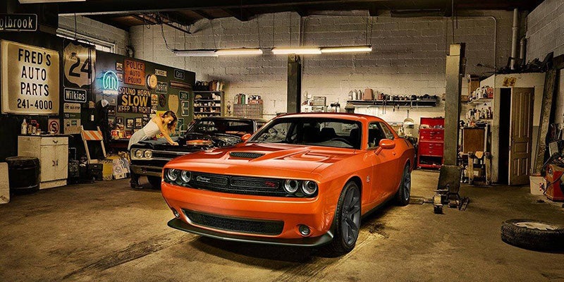 A woman works on a vintage car in a garage with an orange Dodge Challenger in the foreground.