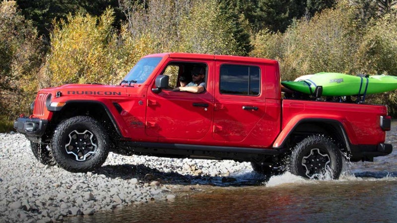 Red Jeep Gladiator off-roading through a shallow river, carrying a bright green kayak in its bed.