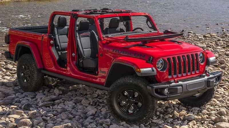 Bright red Jeep Gladiator Rubicon pickup on a rocky riverbank, doors and front windshield removed or folded down.