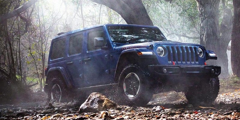 A blue four-door Jeep Wrangler traversing a shallow, rocky stream in a misty forest.
