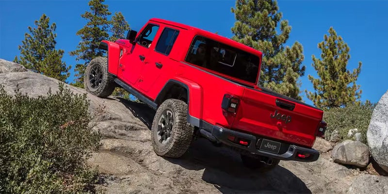 Red Jeep Gladiator truck ascending a rocky off-road trail under a clear blue sky.