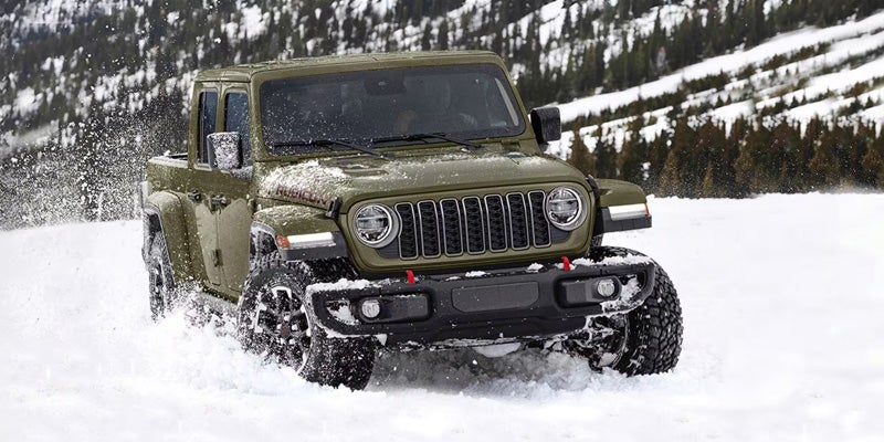 A green Jeep Gladiator Rubicon pickup truck drives through deep snow, kicking up powder, in a wintry forest.