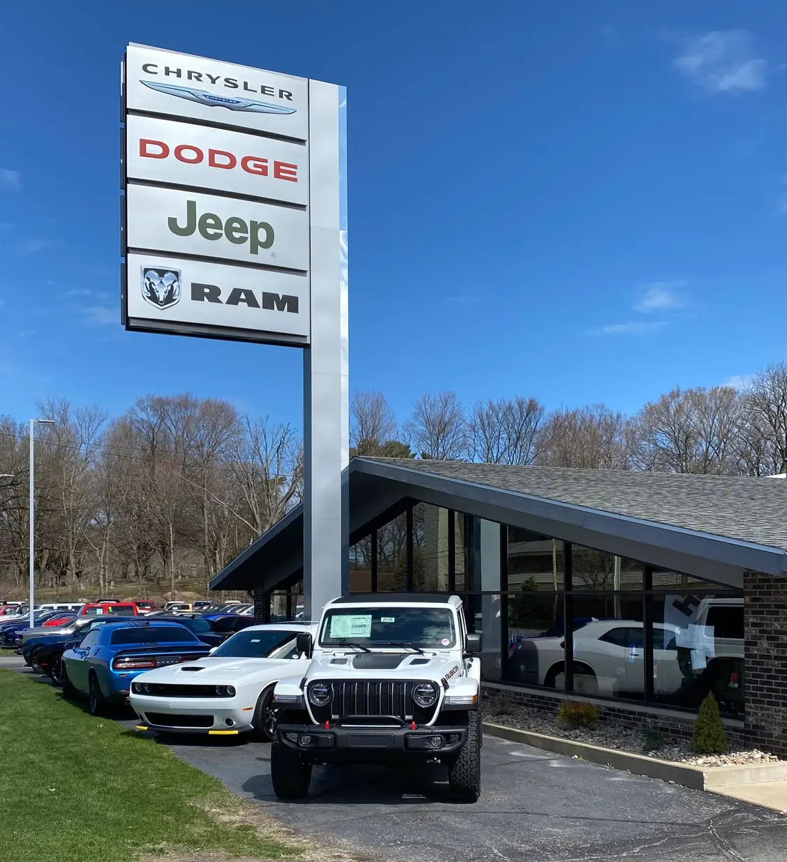 A white Jeep parked in front of a car dealership featuring a Chrysler, Dodge, Jeep, and RAM sign.