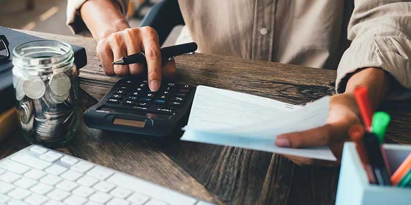 Close-up of a person's hands using a calculator, a jar of coins, and reviewing documents on a wooden desk.